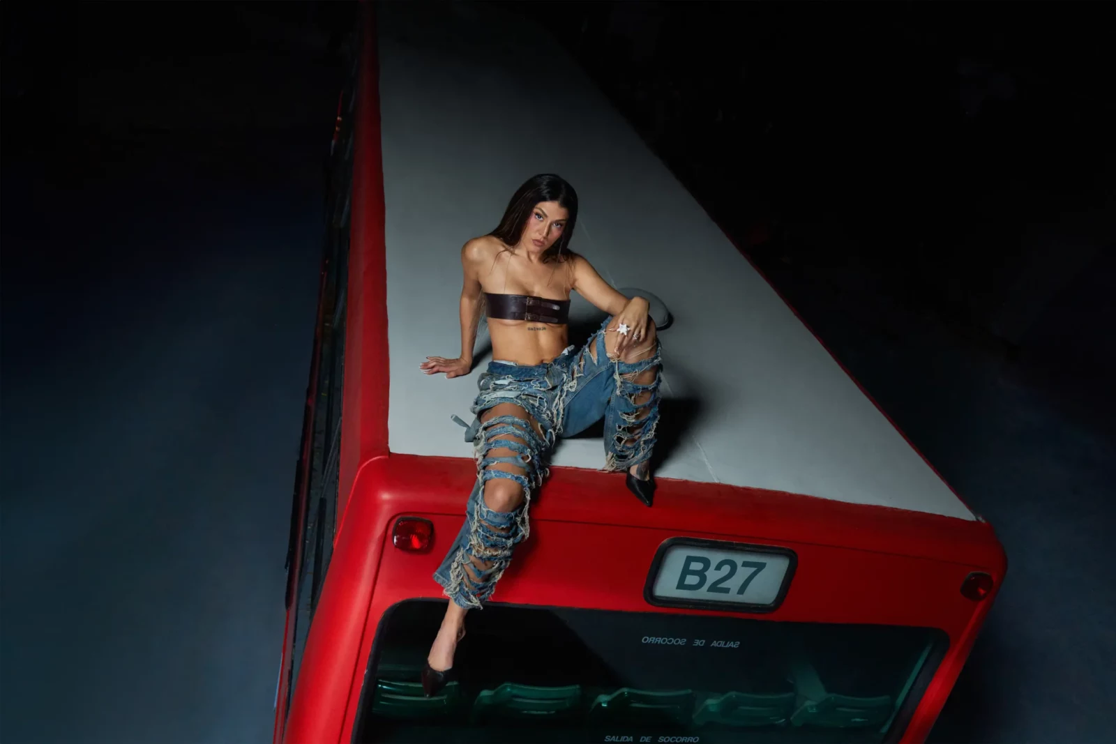 Queralt Lahoz seated on the front of a red bus in patterned blue jeans and a black top.