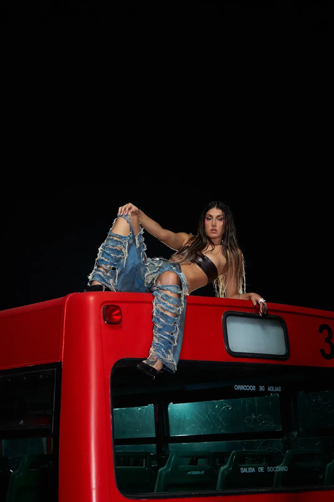 Queralt Lahoz in jeans and a black crop top posing on the roof of a red city bus at night.