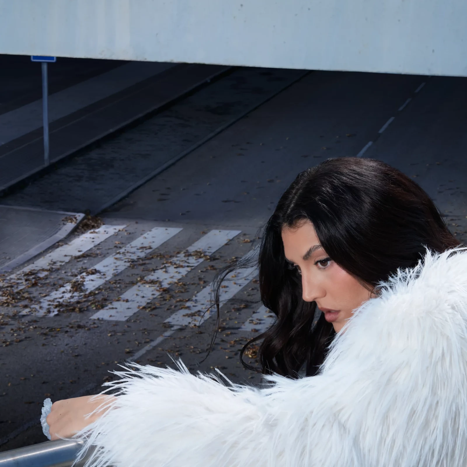 Side portrait of Queralt Lahoz in a white faux-fur coat above a worn pedestrian crossing.