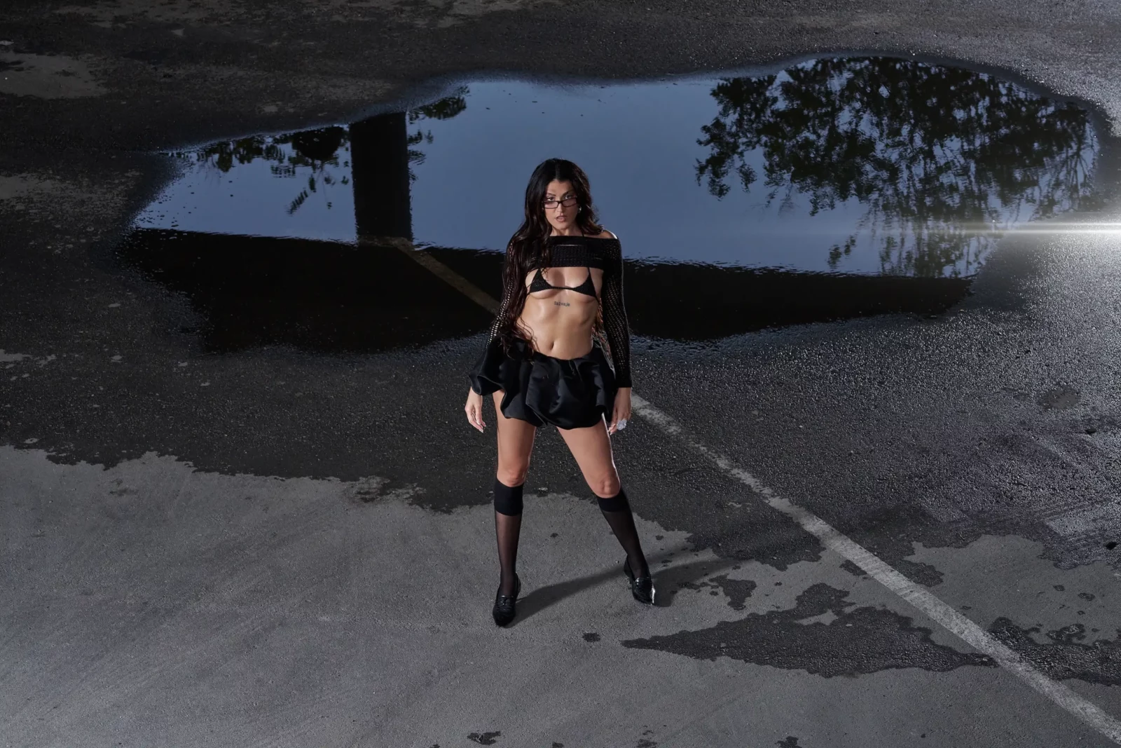 Queralt Lahoz in a black two-piece outfit and tall boots standing beside a reflective puddle at dusk.