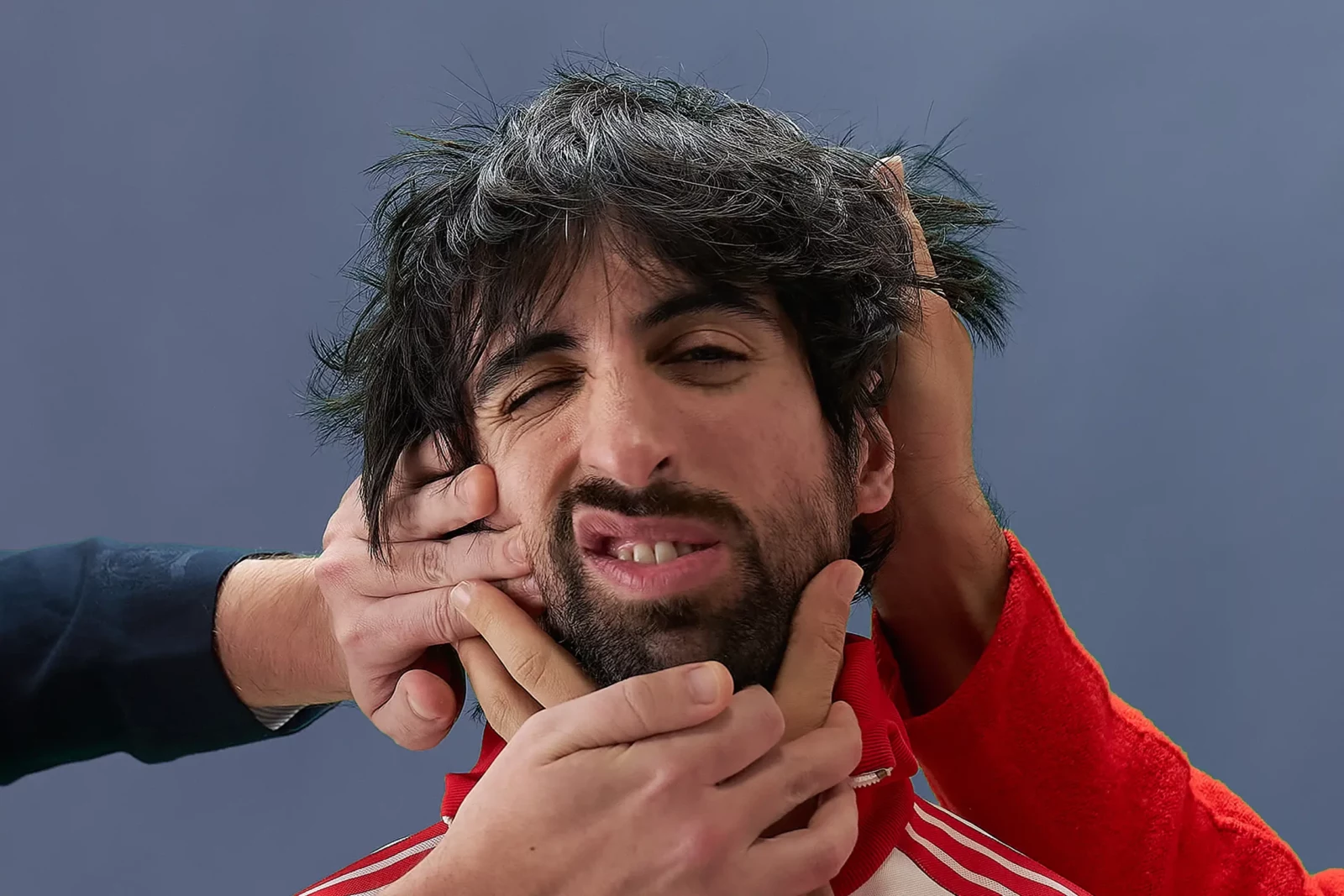Portrait of a dark-haired Cupido musician as hands press his cheeks against a pale blue backdrop.