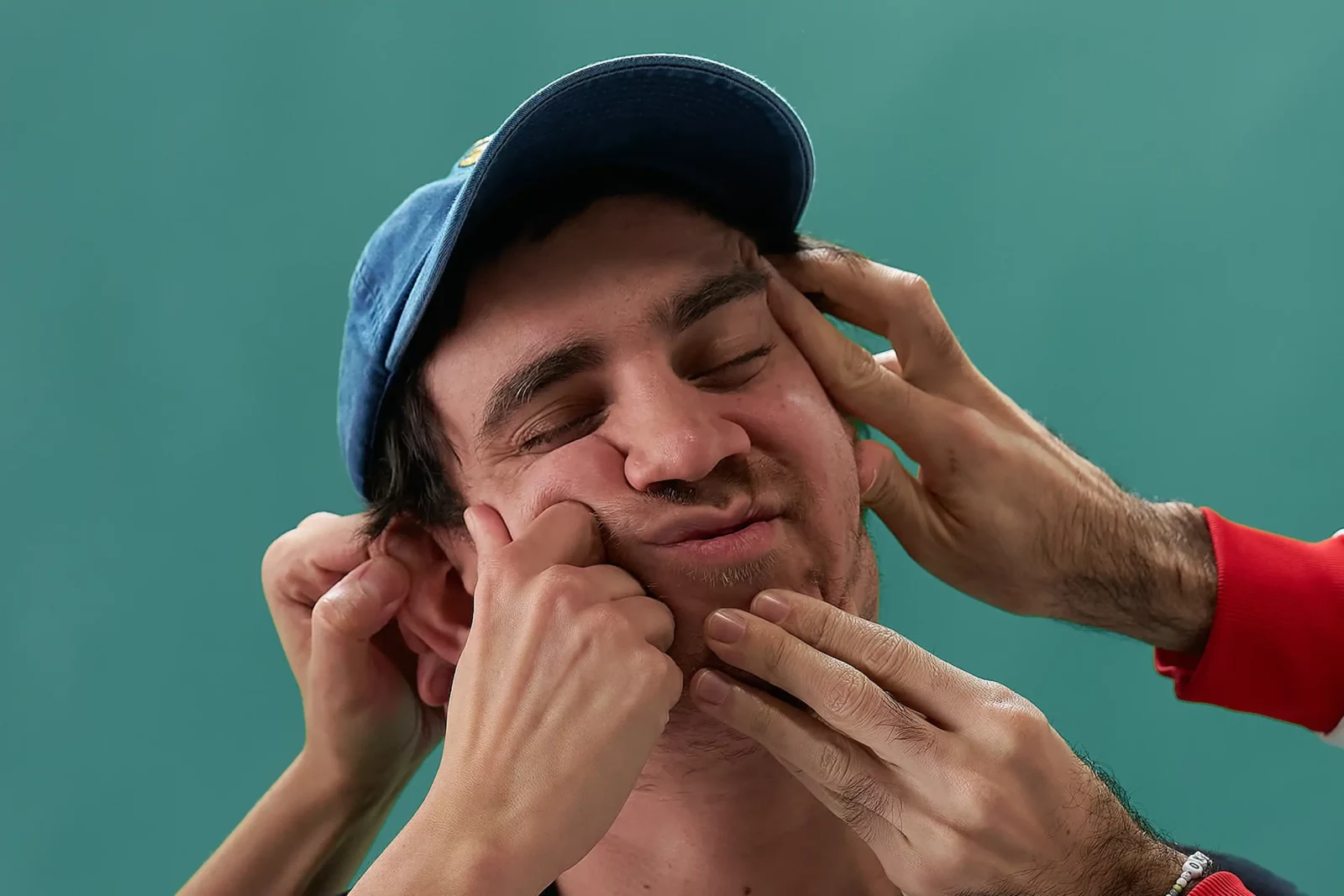 Cupido band member wearing a baseball cap with hands squeezing his face against a teal backdrop.