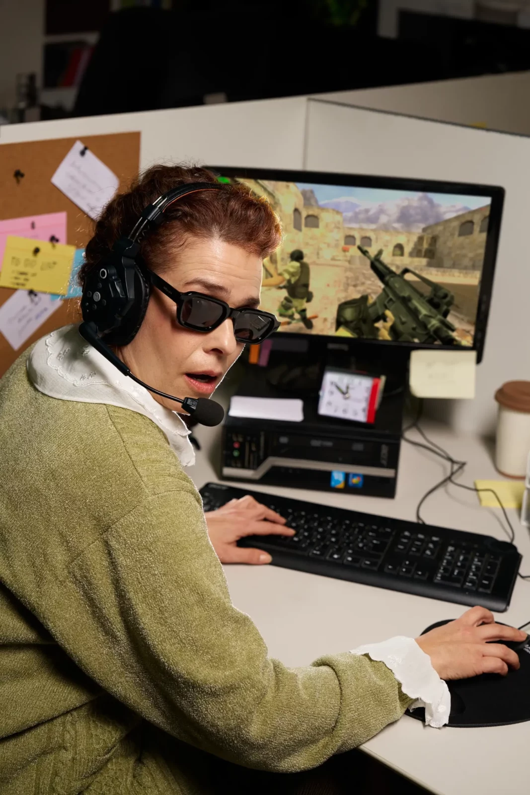 Man in sunglasses seated at a desk using a computer in a busy retro office.