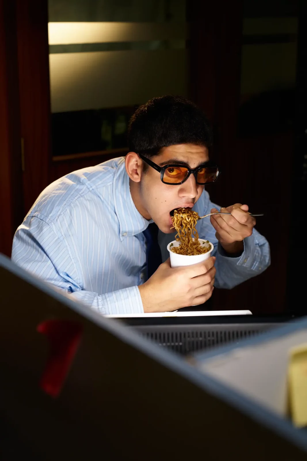 Man wearing sunglasses and a light blue shirt leaning across an office desk with a drink.