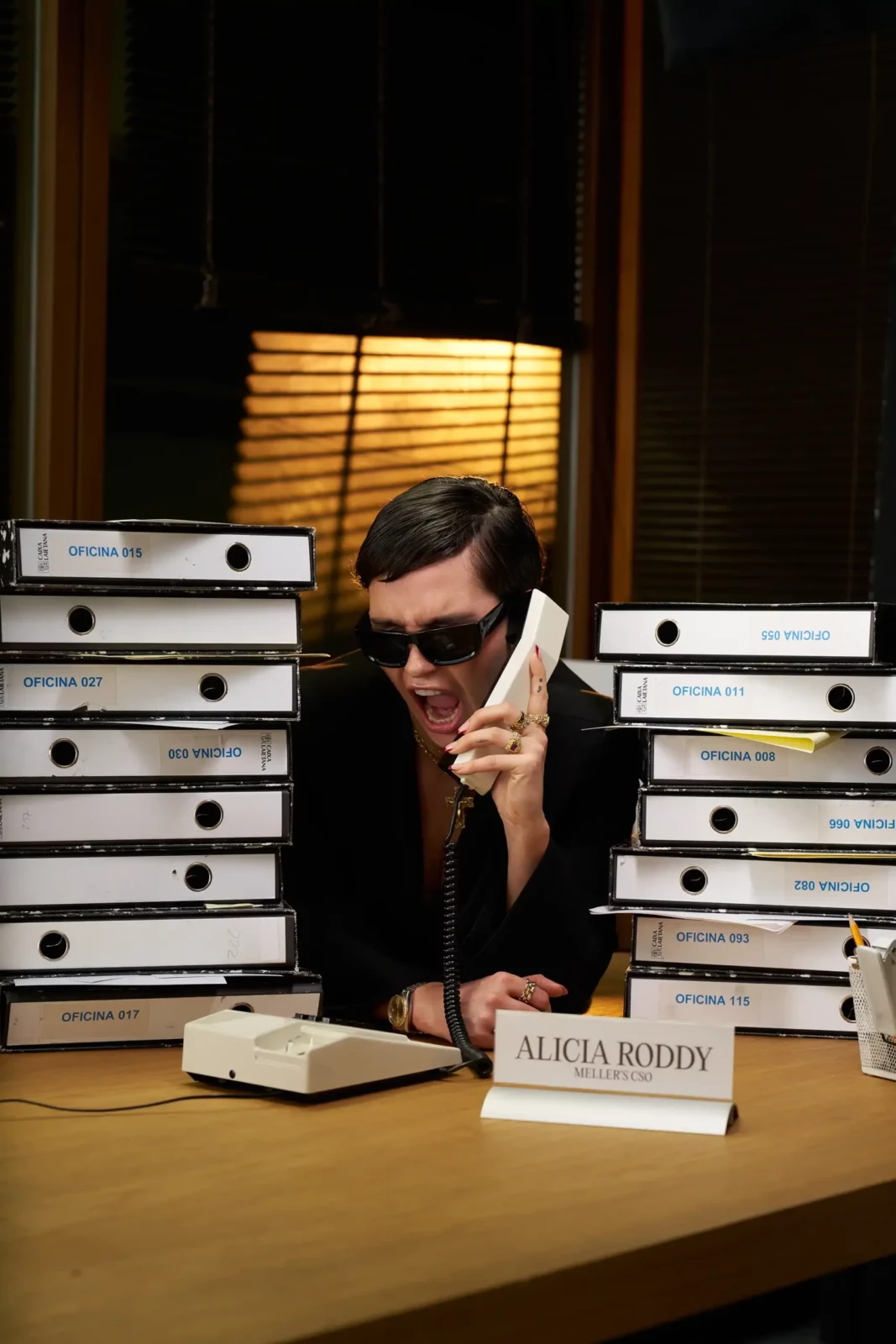 Woman in black sunglasses seated at an office desk surrounded by stacks of white boxes.