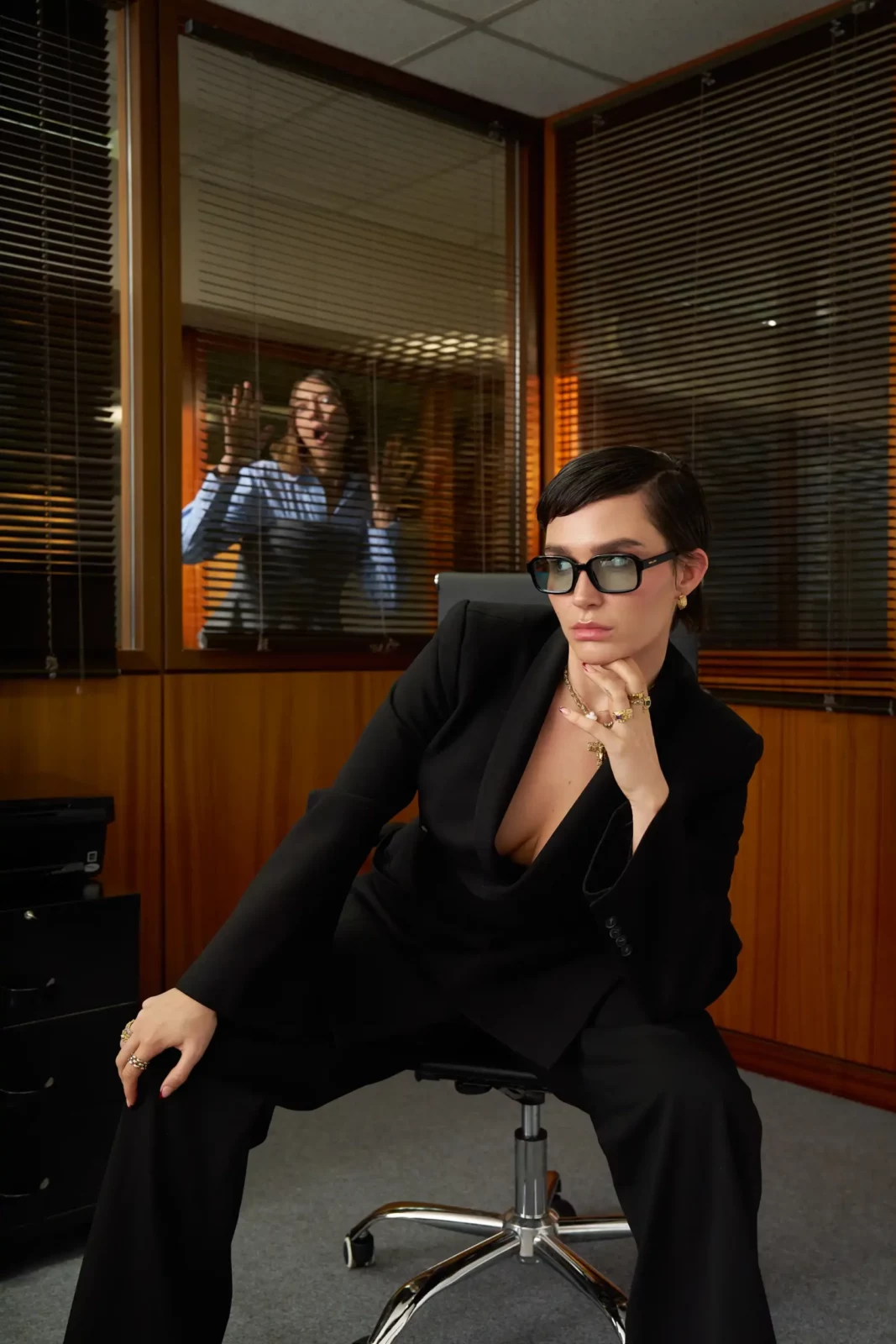 Model dressed in black and sunglasses seated at a desk in a wood-paneled office.