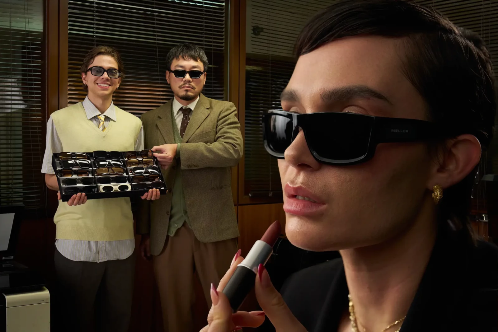 Close-up of a woman in black sunglasses in the foreground of an office scene with colleagues behind her.