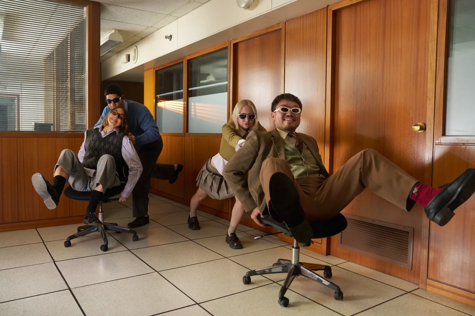 Three men wearing sunglasses and formal clothing seated in a retro office interior.