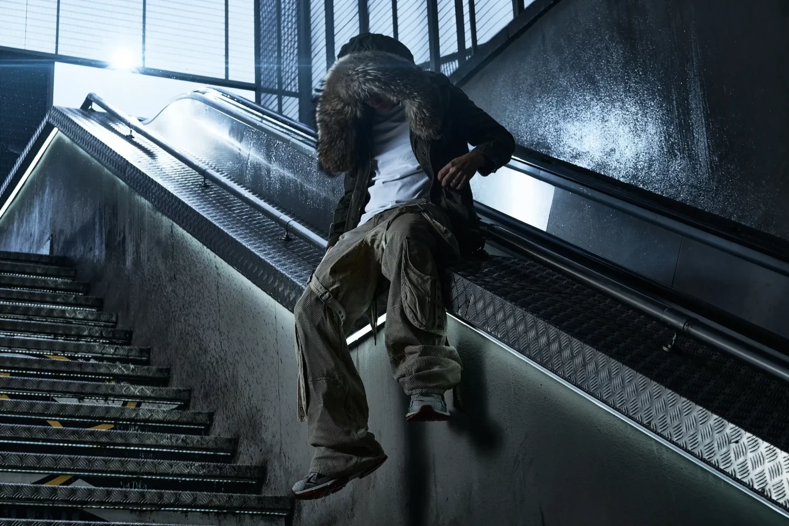 Model in baggy trousers and a hooded jacket moving along a concrete stairwell and escalator.