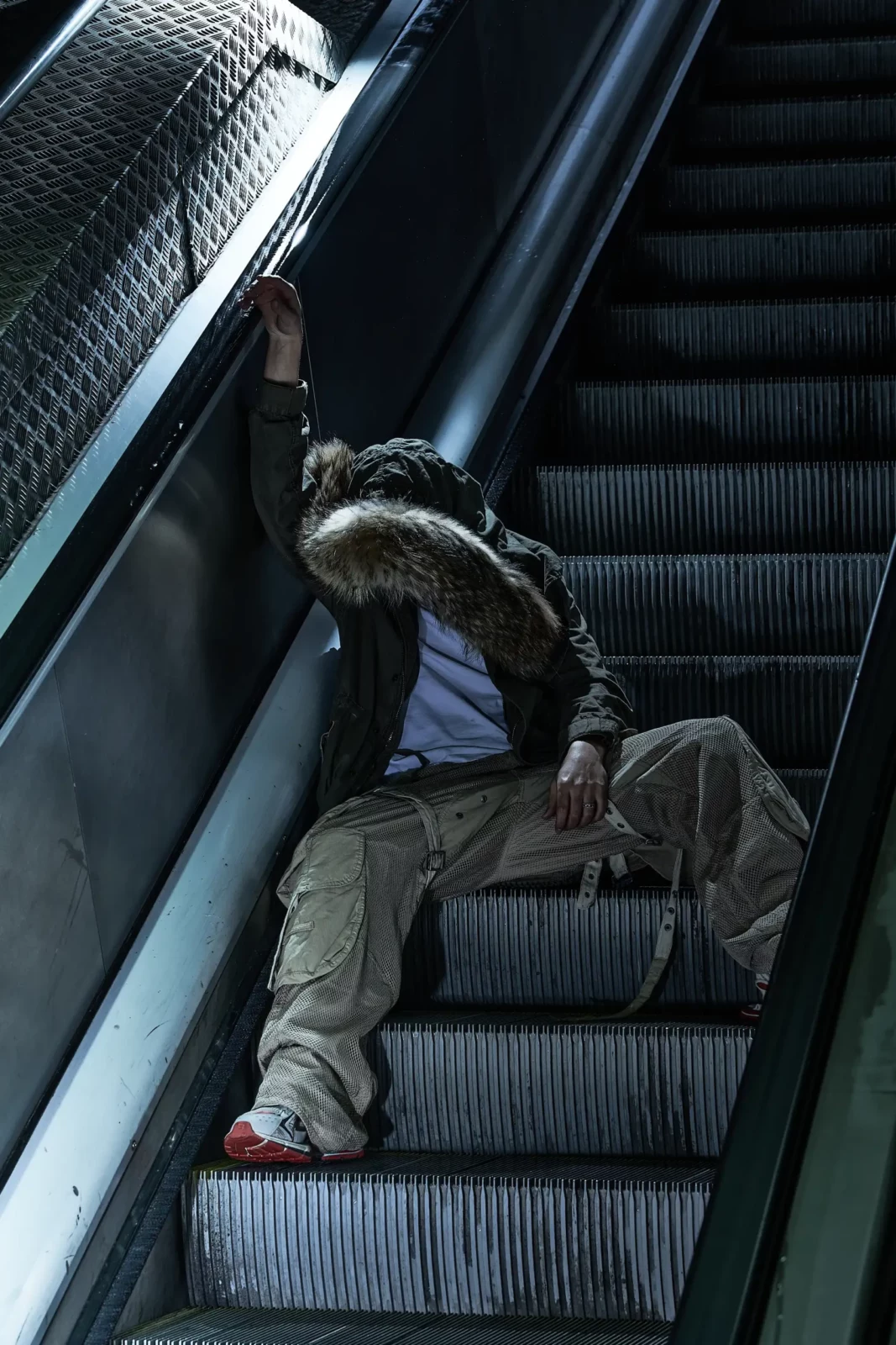 Model wearing a heavy hooded jacket and loose trousers seated on a moving escalator.