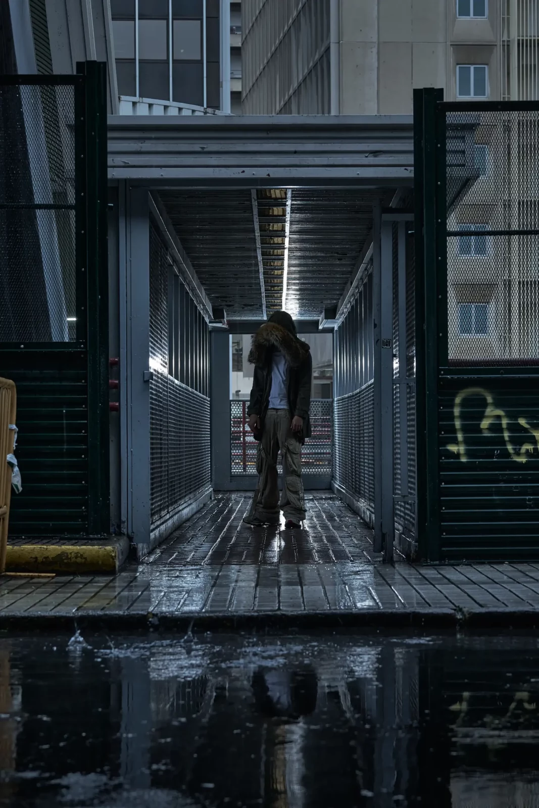 Full-length photograph of a hooded model standing in an industrial covered pedestrian walkway.