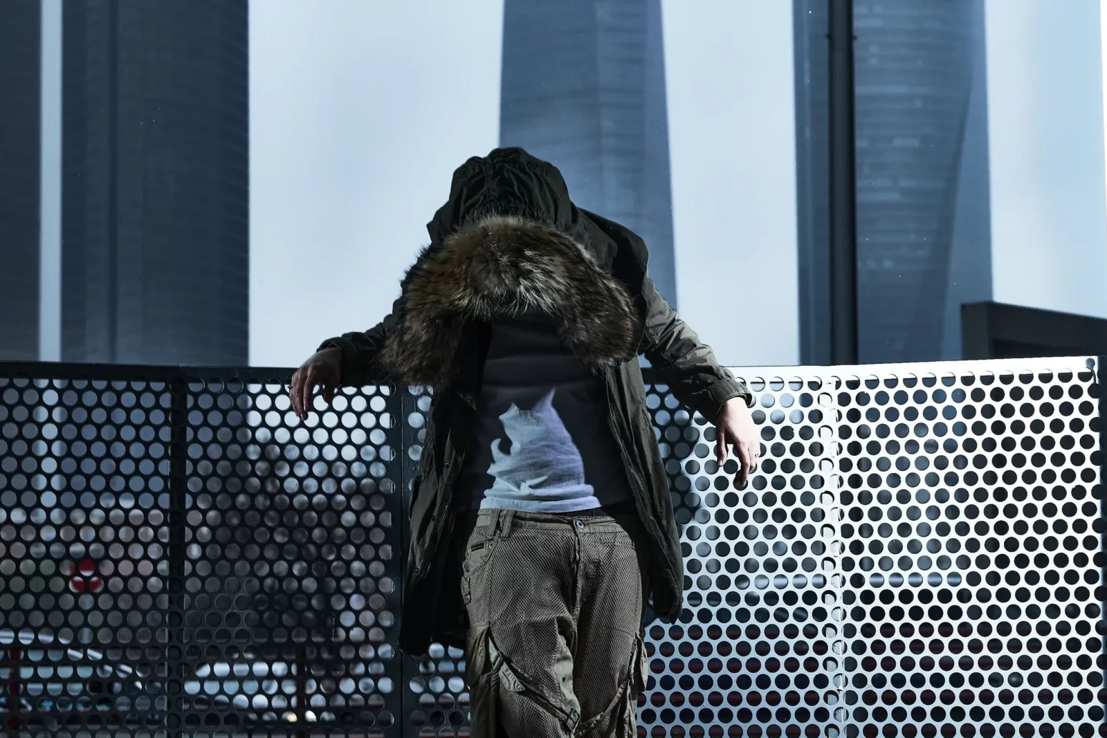 Rear view of a model in a parka with an oversized furry hood leaning against perforated metal railing.
