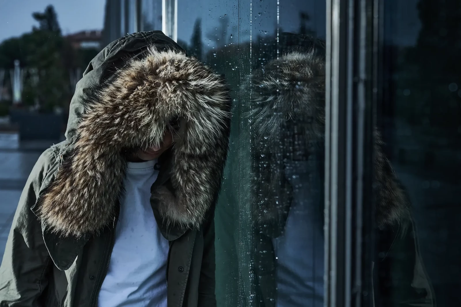 Model wearing a shaggy fur-trimmed hood standing next to reflective glass with an urban reflection.