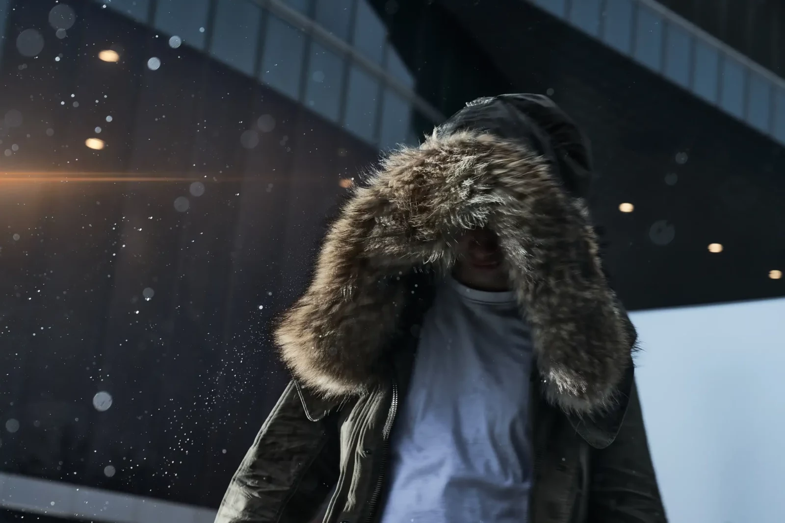 Close-up of a model in a brown parka with a large fur-trimmed hood beside a modern glass facade.