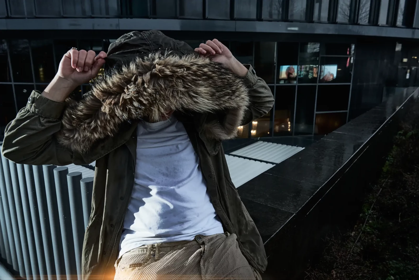 Fashion model adjusting an oversized fur-lined hood while wearing a white T-shirt and dark jacket outdoors.