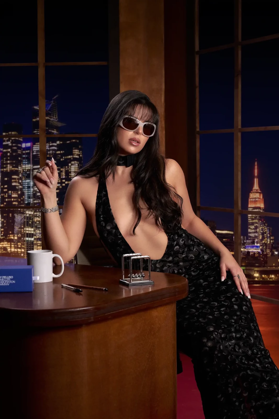Nathy Peluso seated behind a wooden late-night-show desk wearing white sunglasses and a deep V-neck black dress.
