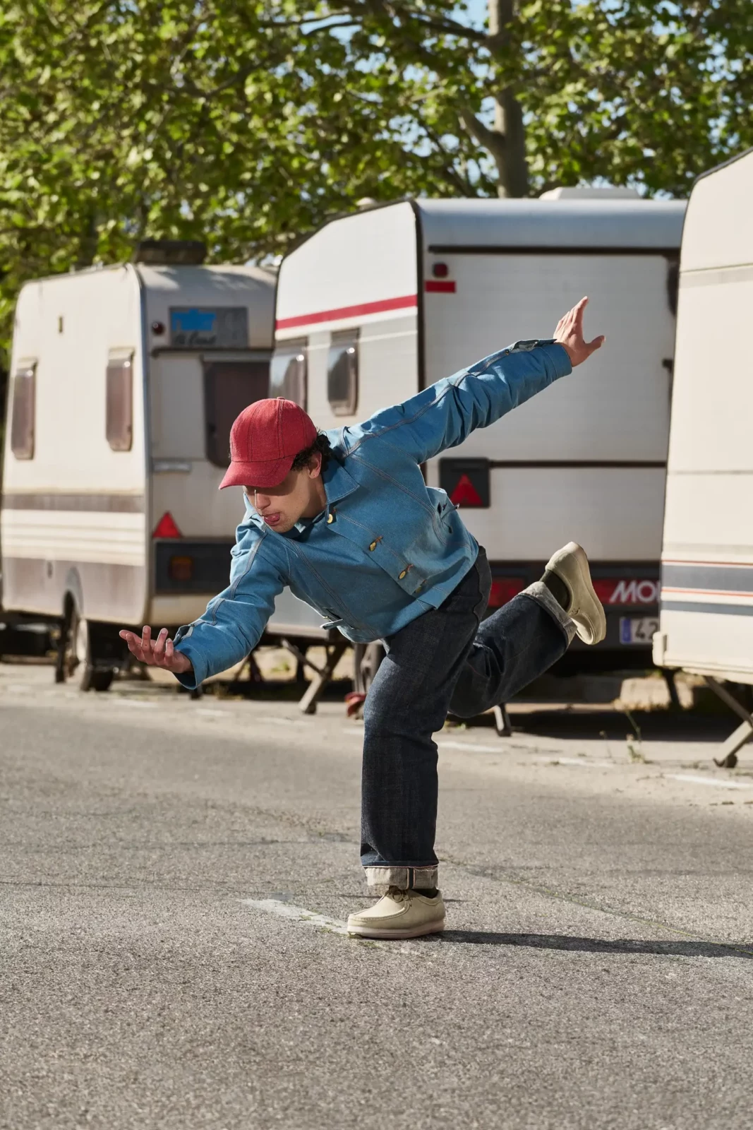 Model in a navy jacket and red skirt leaning sideways on a paved path near a caravan.