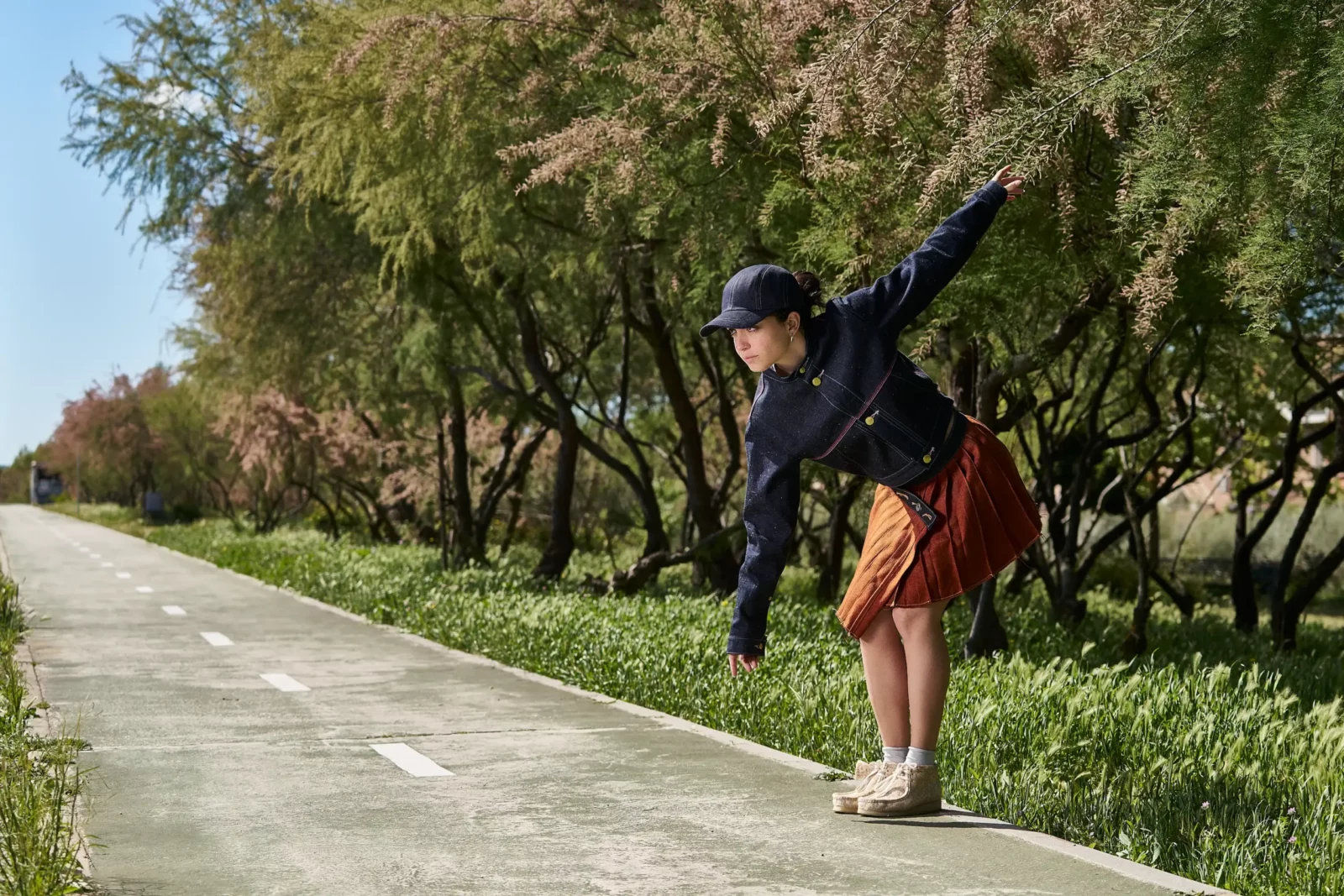Fashion model in a cap and red skirt balancing on a sunlit country road.