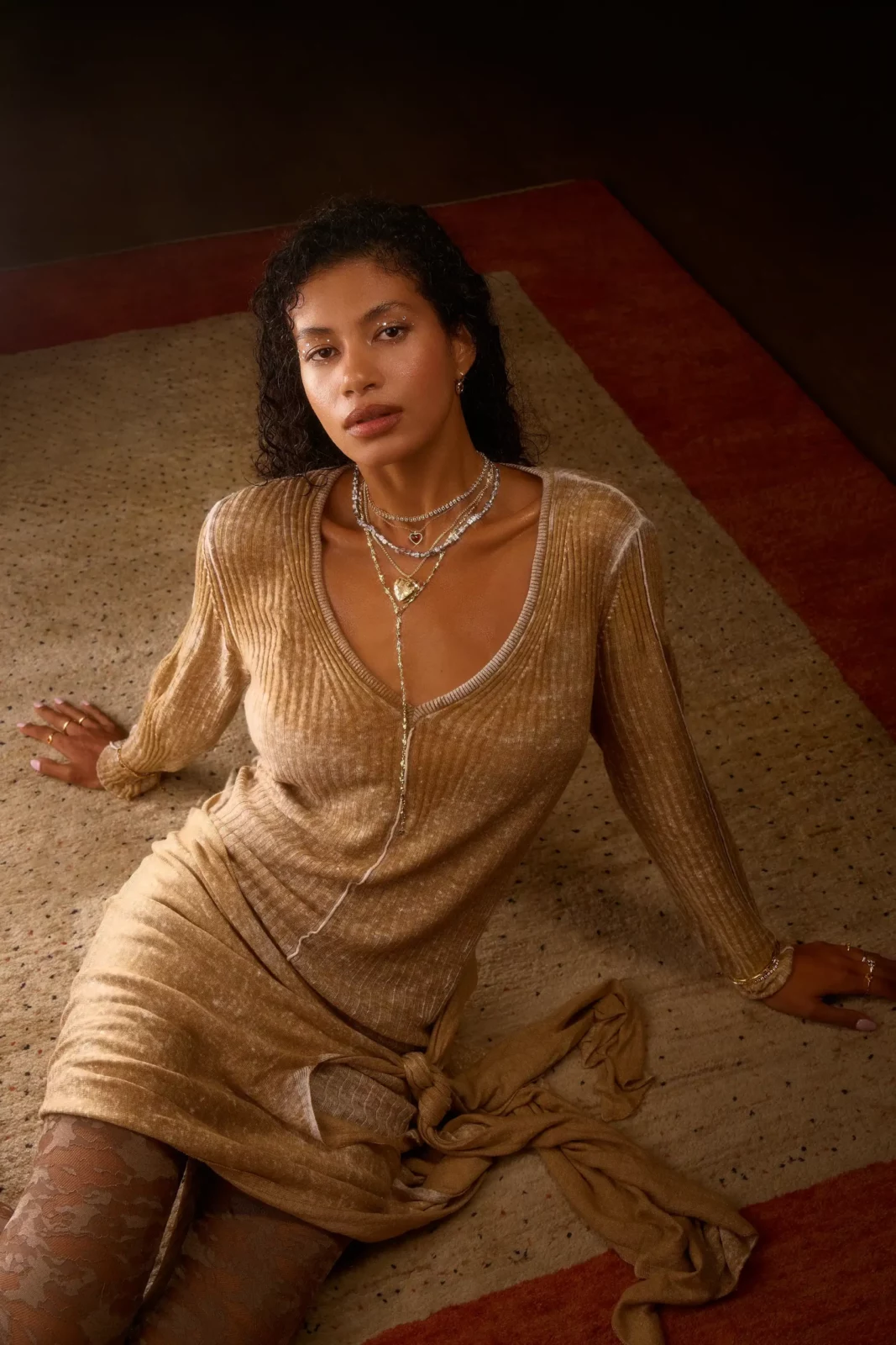 Seated model wearing a shimmering gold dress and layered jewelry in a warm sculptural interior.