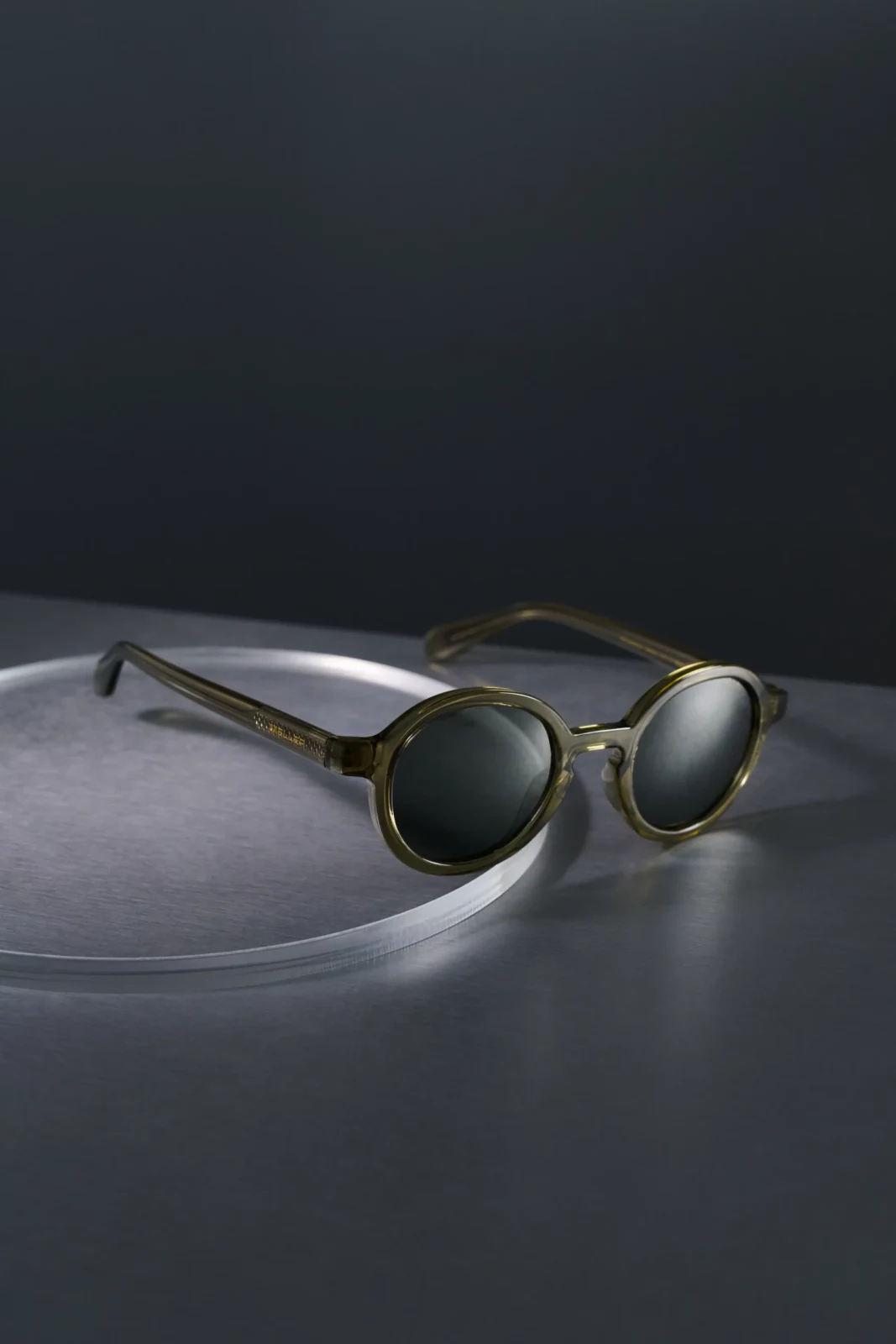 Still-life photograph of green-tinted sunglasses resting on a dark curved surface.