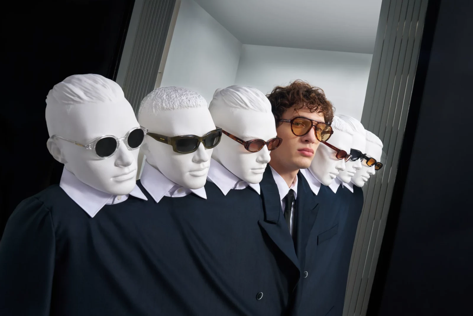 Man in a suit and sunglasses walking alongside figures wearing white fabric hoods in an office corridor.