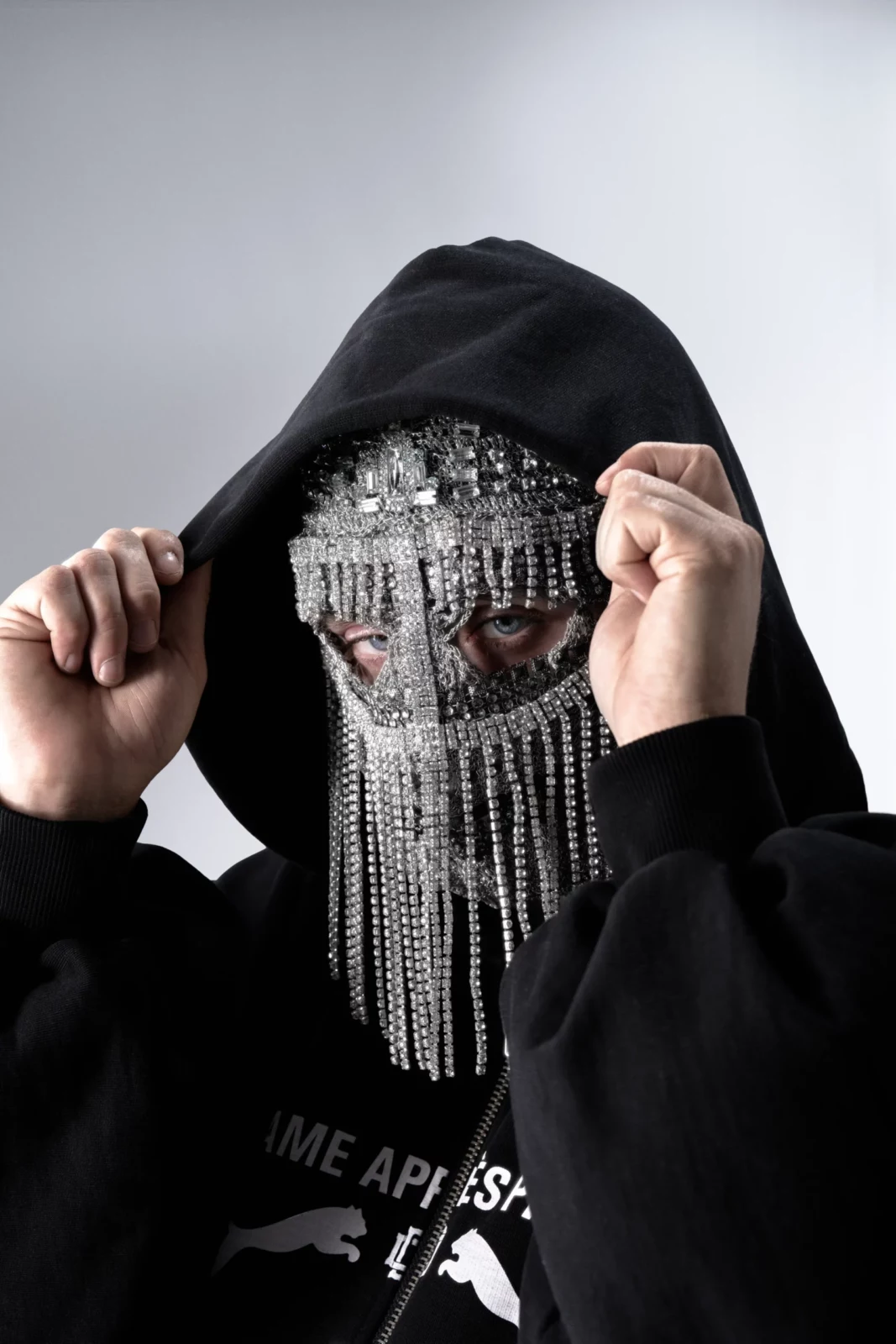 Close-up portrait of a hooded figure lifting a black sweatshirt to reveal long dangling silver face chains.