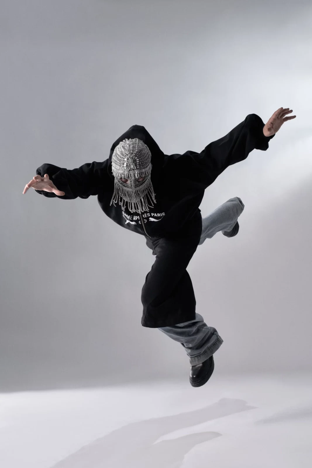 Masked performer in oversized dark clothing jumping against a minimalist gray studio background.