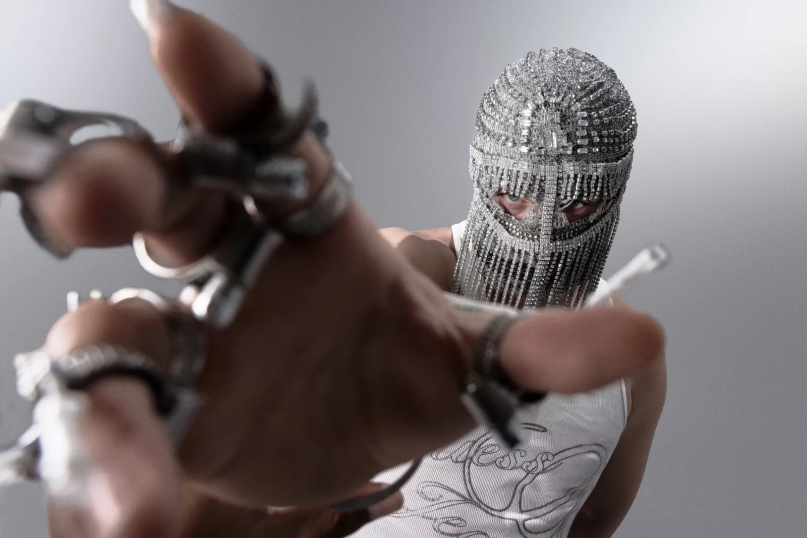 Close-up of a hand with elaborate metal finger rings reaching toward the camera in front of a masked face.