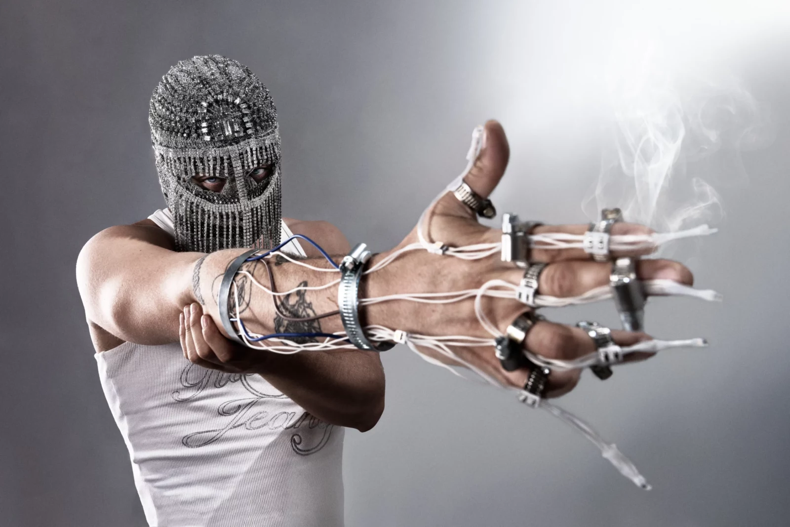 Masked performer in a white tank top stretching both hands forward through sculptural metal finger jewelry.