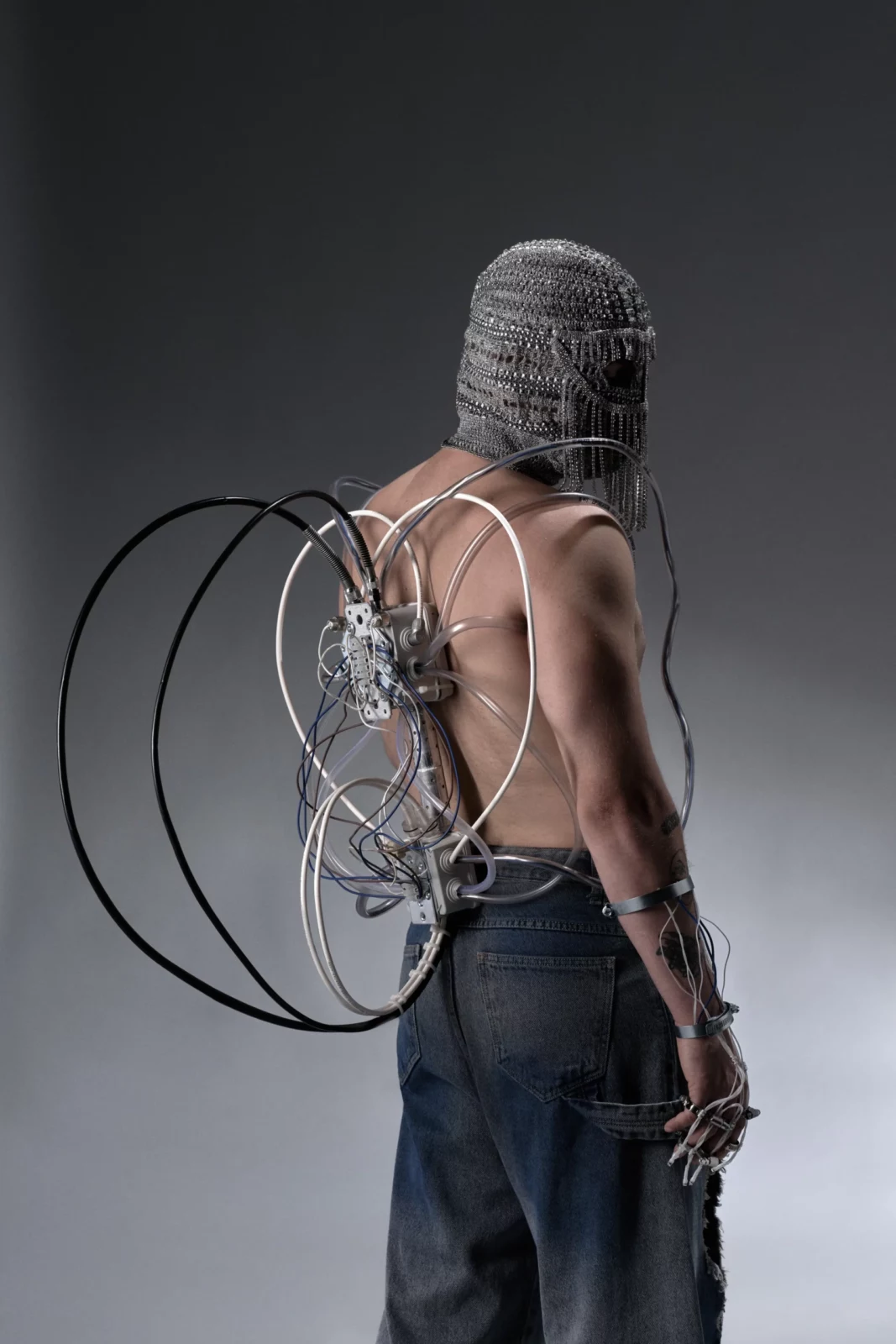 Shirtless model with a silver face covering and a circular metal sculpture attached to the back.
