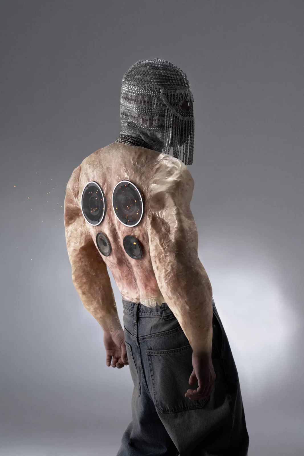Rear three-quarter portrait of a shirtless performer in a silver mesh hood showing circular markings on the back.