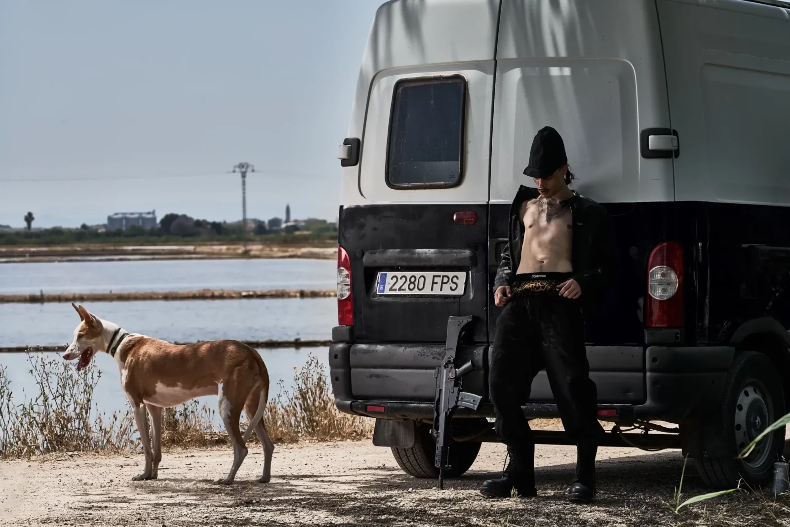 Fashion model standing beside a parked truck and a dog near a shallow coastal wetland.