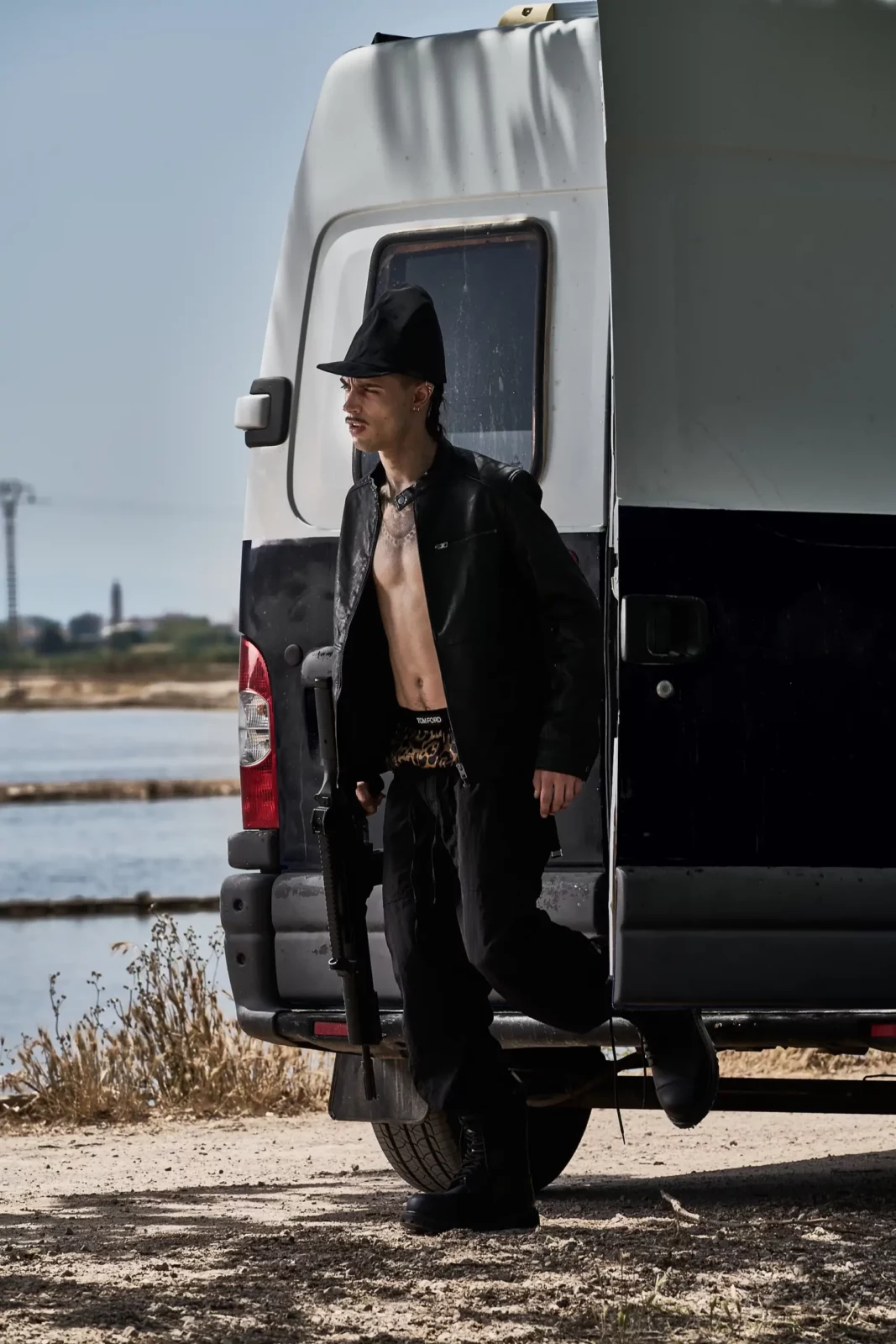 Model wearing an open black jacket and cap leaning beside a white truck.