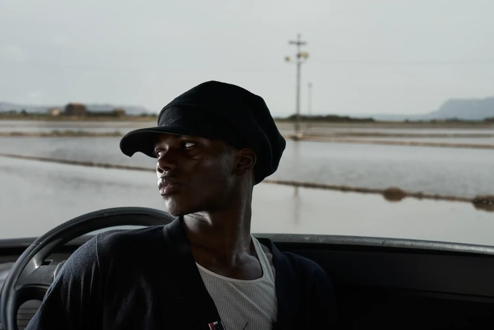Portrait of a model in a black cap sitting inside a vehicle against a coastal landscape.