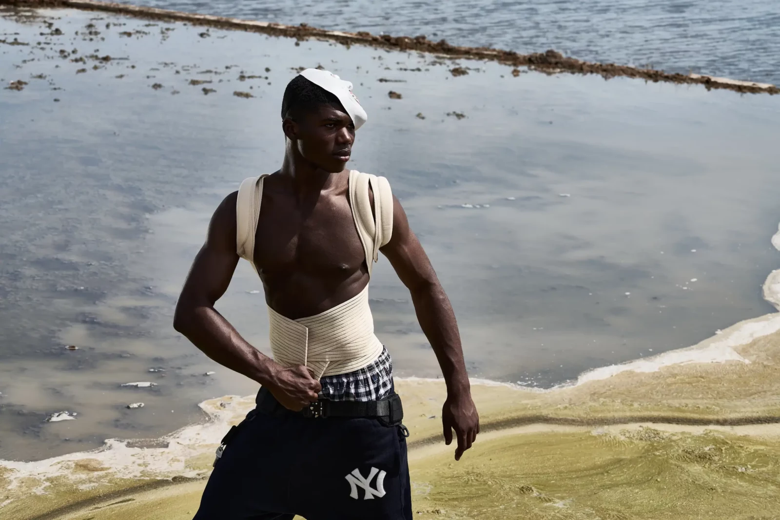 Model wearing a white tank top and dark trousers standing at the edge of shallow water.