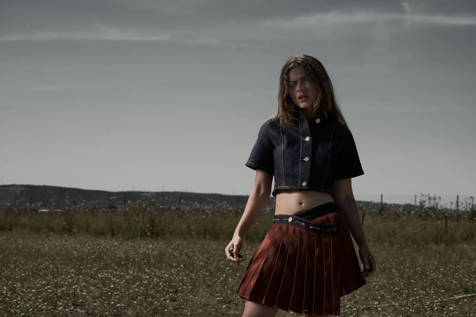 Young woman in a cropped top and pleated red skirt standing in an open field.