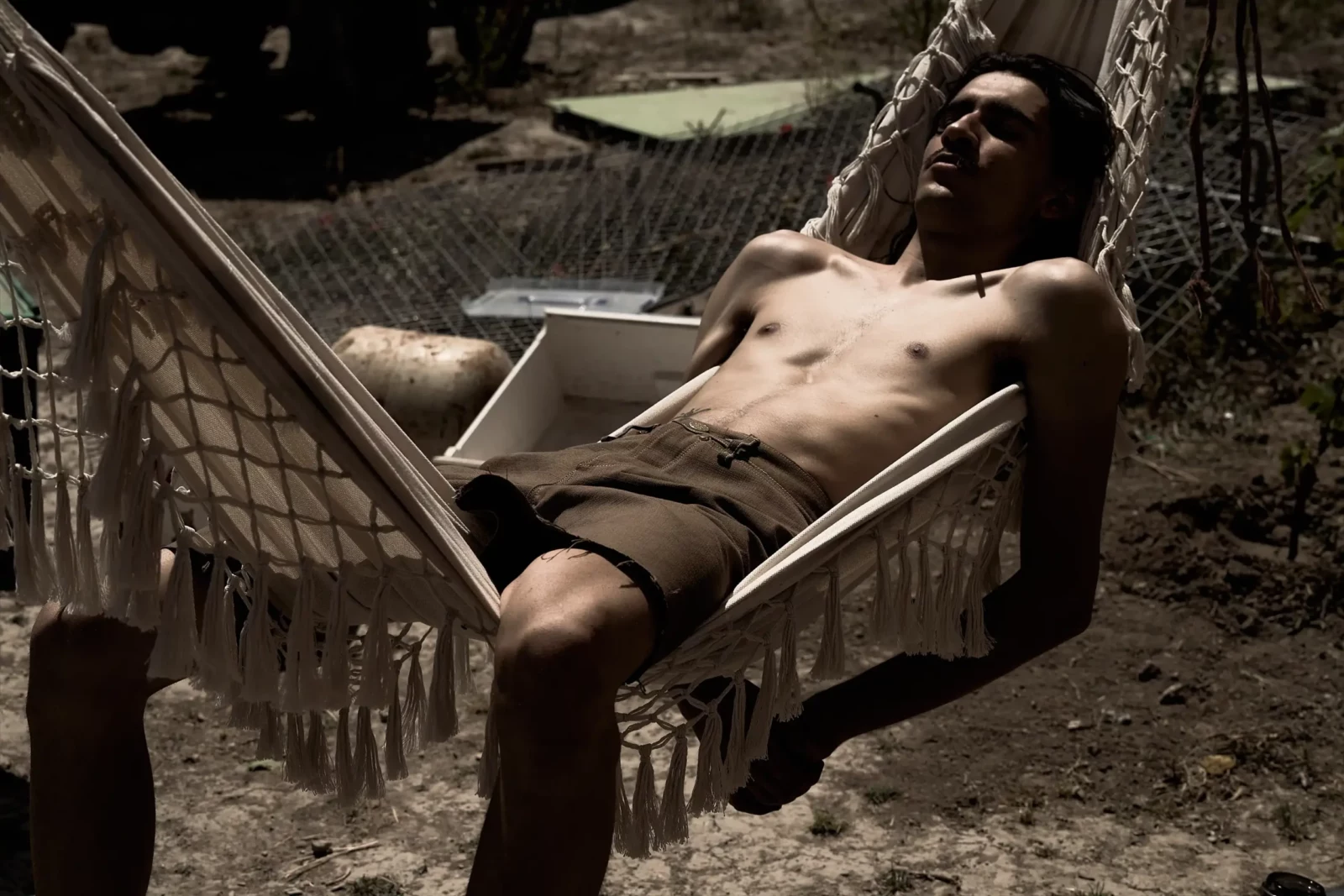 Shirtless young man reclining in a suspended hammock outdoors in the summer heat.