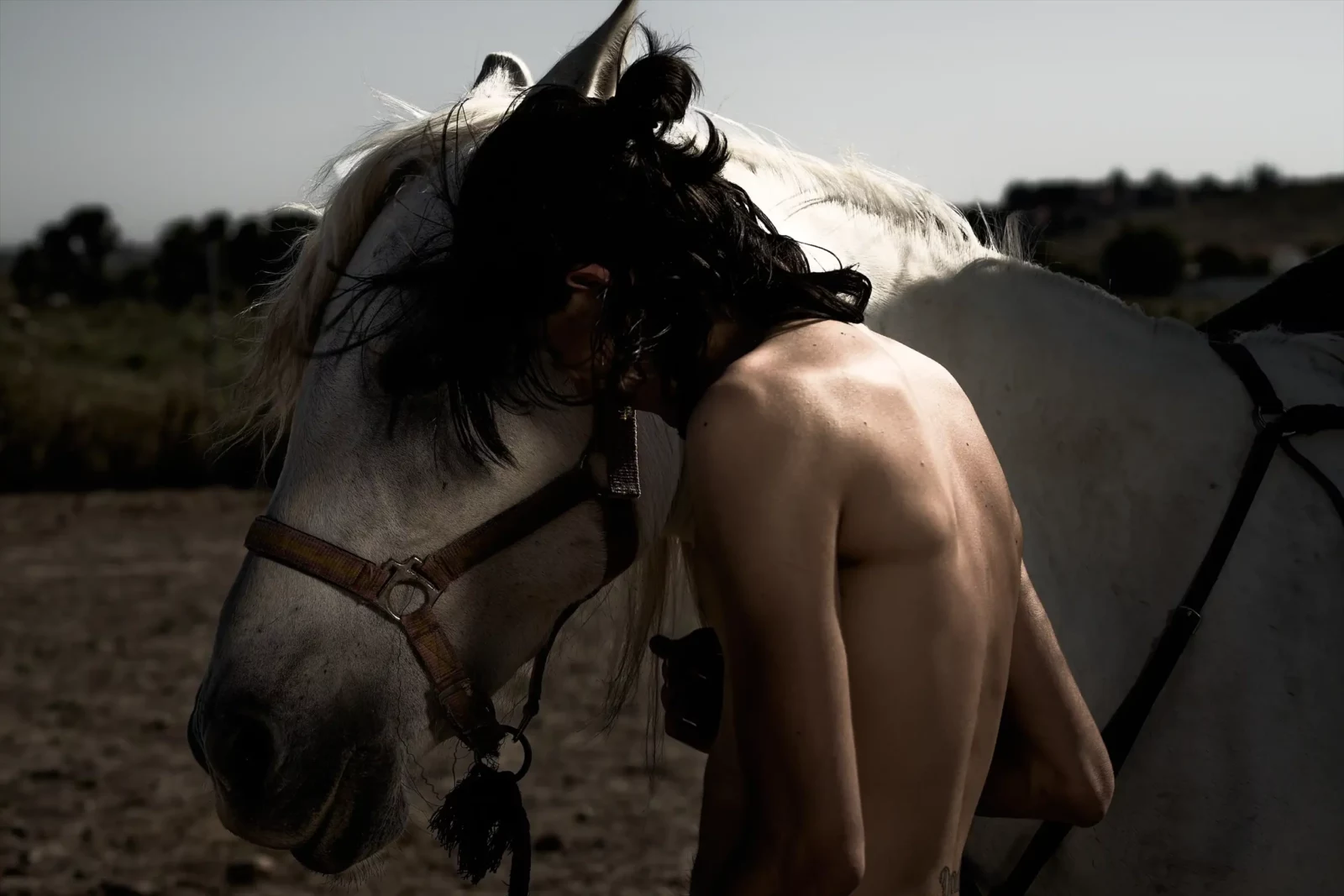 Shirtless young man standing beside a white horse in a rustic outdoor setting.