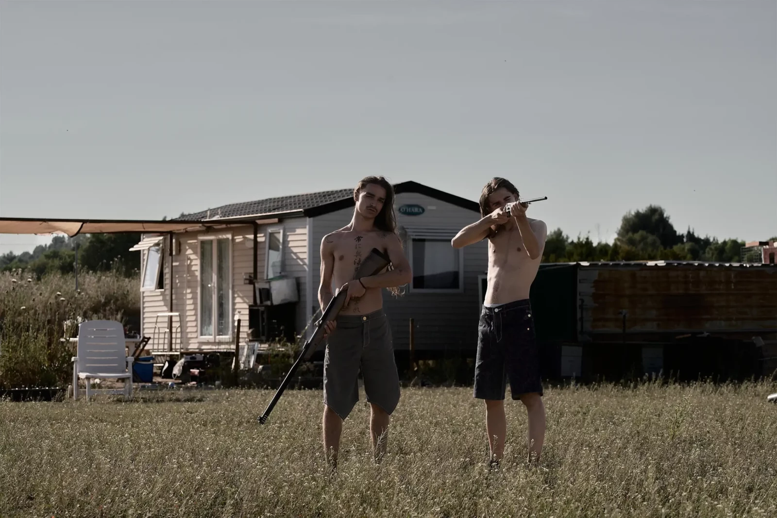 Two shirtless models holding a branch outside a caravan in a grassy landscape.