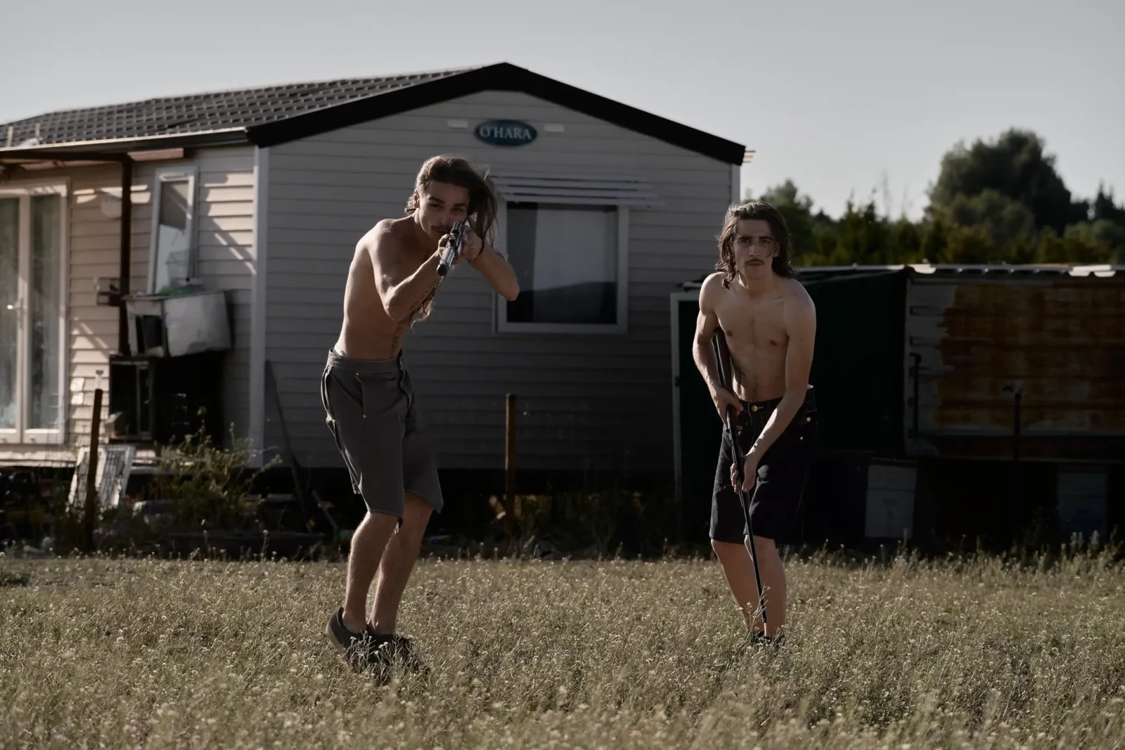 Two young men running away from a caravan through a sunlit grassy field.