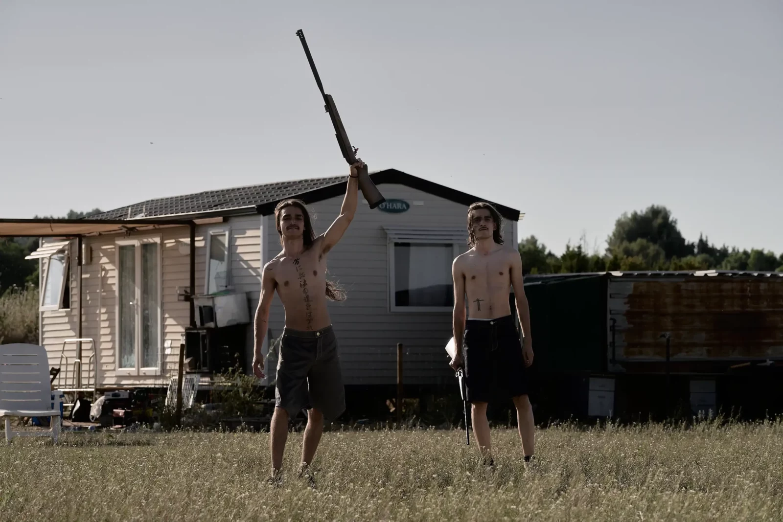 Two young men standing with long branches in a dry field beside a small caravan.