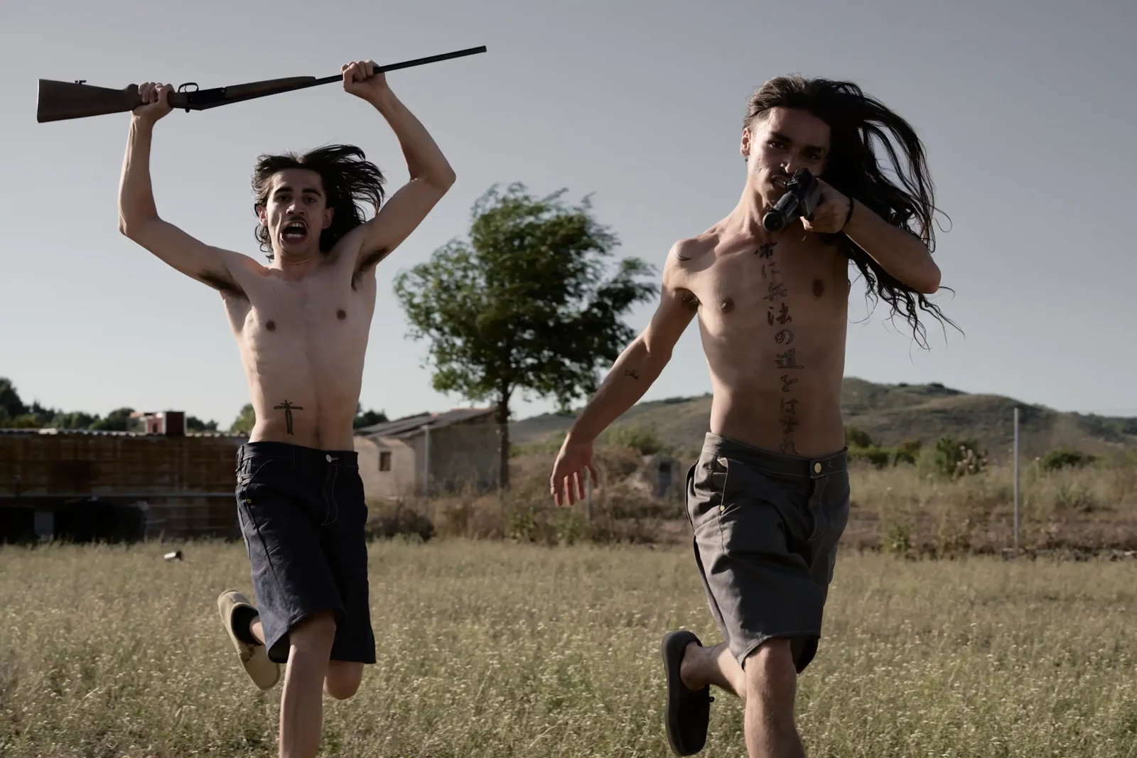 Two shirtless men running through an open field carrying a long branch above their heads.
