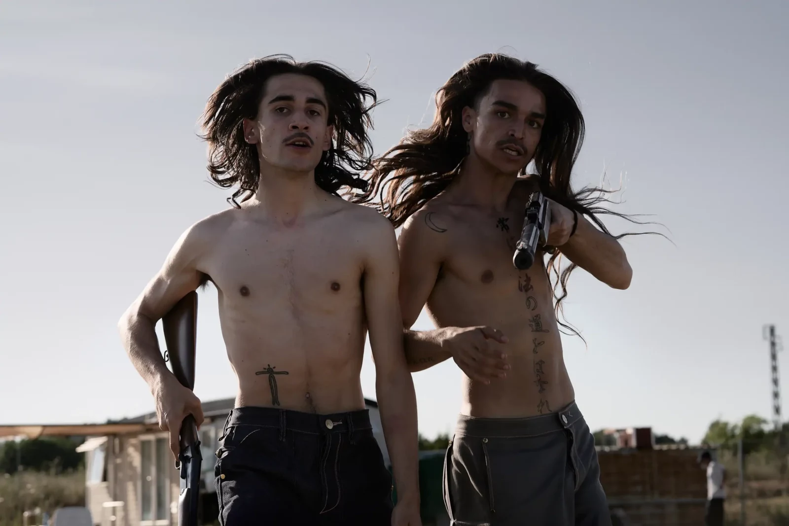 Two shirtless men running through a dry grassy field with their hair blowing in the wind.