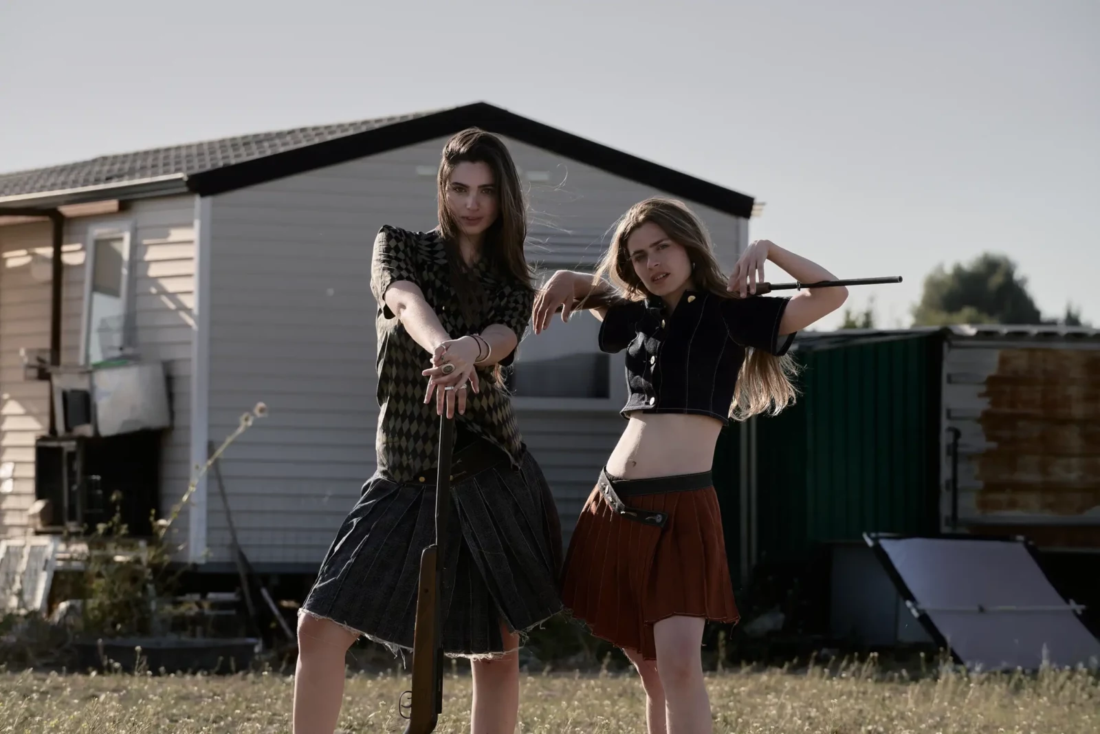 Two fashion models standing with branches beside a caravan in a sunlit field.