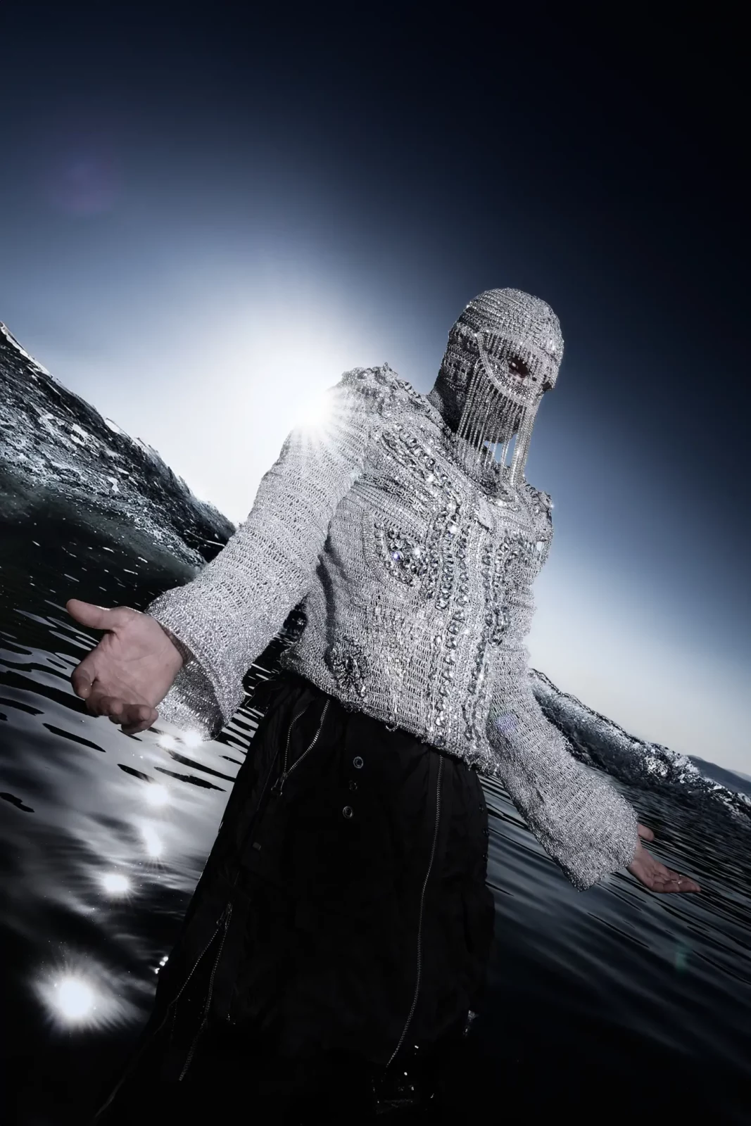 Performer in a reflective mesh hood and textured white top leaning on a rock beside dark water.
