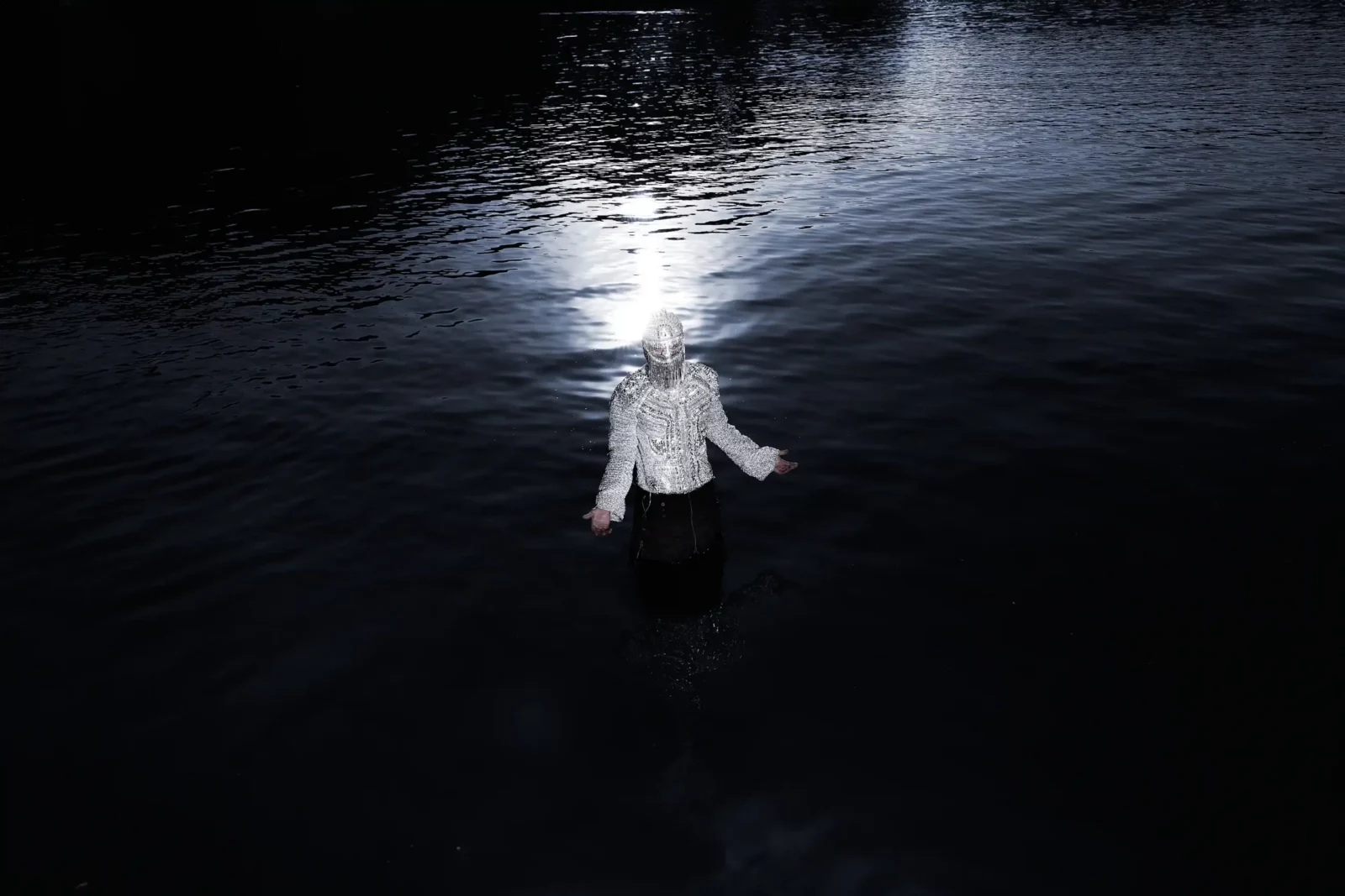 Wide nighttime photograph of a silver-clad figure standing in black water with a bright reflection behind them.