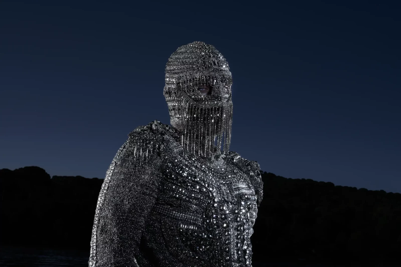 Nighttime portrait of a performer in a reflective metallic mesh hood and silver jacket against dark hills.