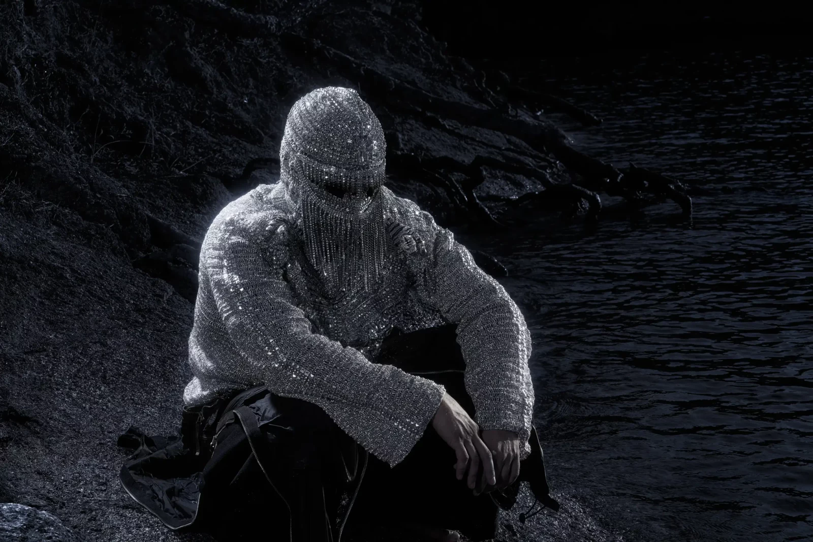 Seated figure in a glittering silver face covering and textured top on dark rocky terrain at night.