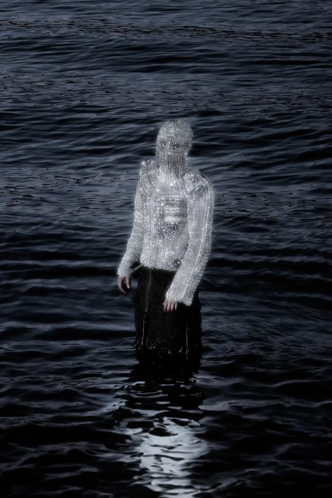 Full-length figure wearing a silver mesh hood and black trousers standing in shallow dark water.