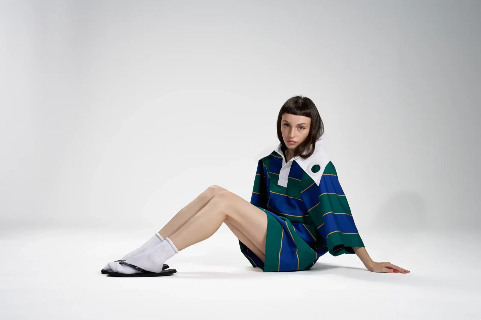 Seated model in an oversized blue and green striped polo shirt, beige shorts and white socks against a white studio backdrop.